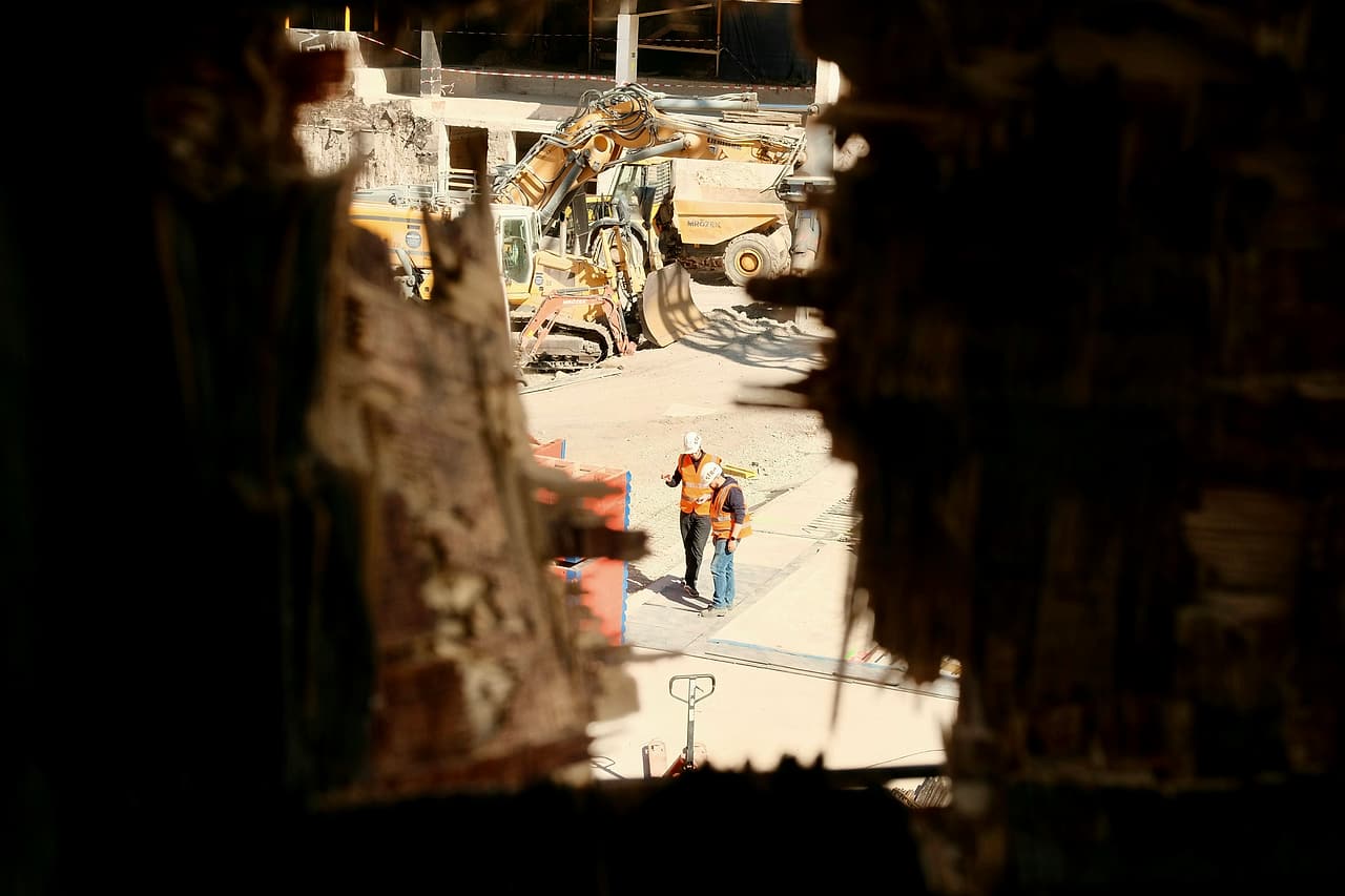 Seen through a gap in a demolished wall, two people in hard hats and high-visibility vests walk and talk across a site.