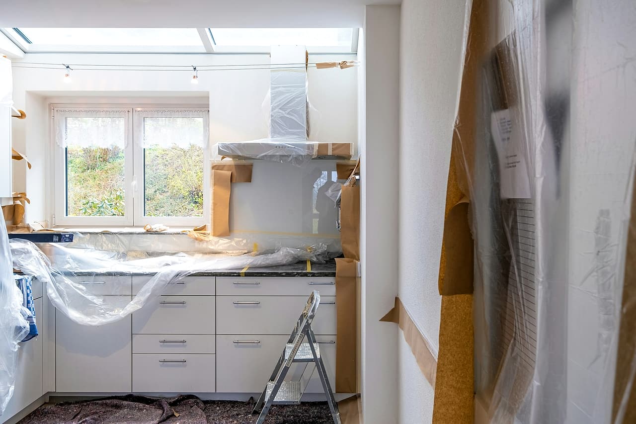 A kitchen under renovation with counters, cabinets and range hood covered in plastic and paper, and a step stool left open.