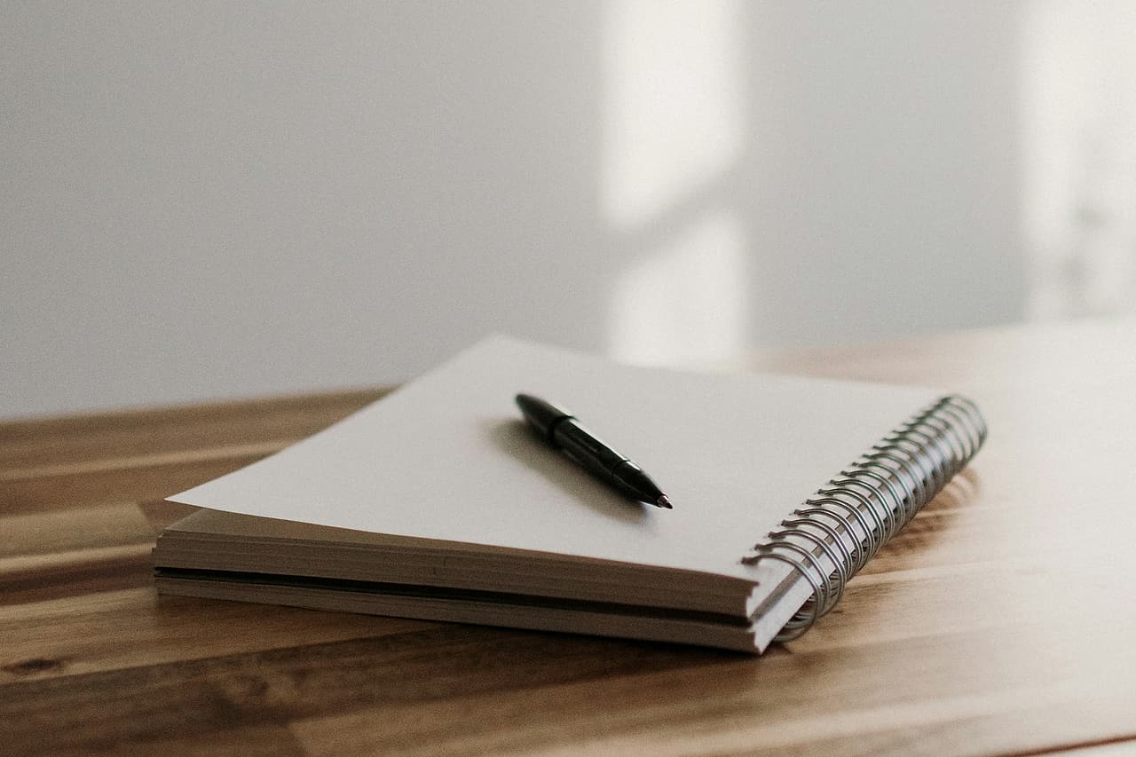 A pen rests on a blank spiral notebook beside a window.