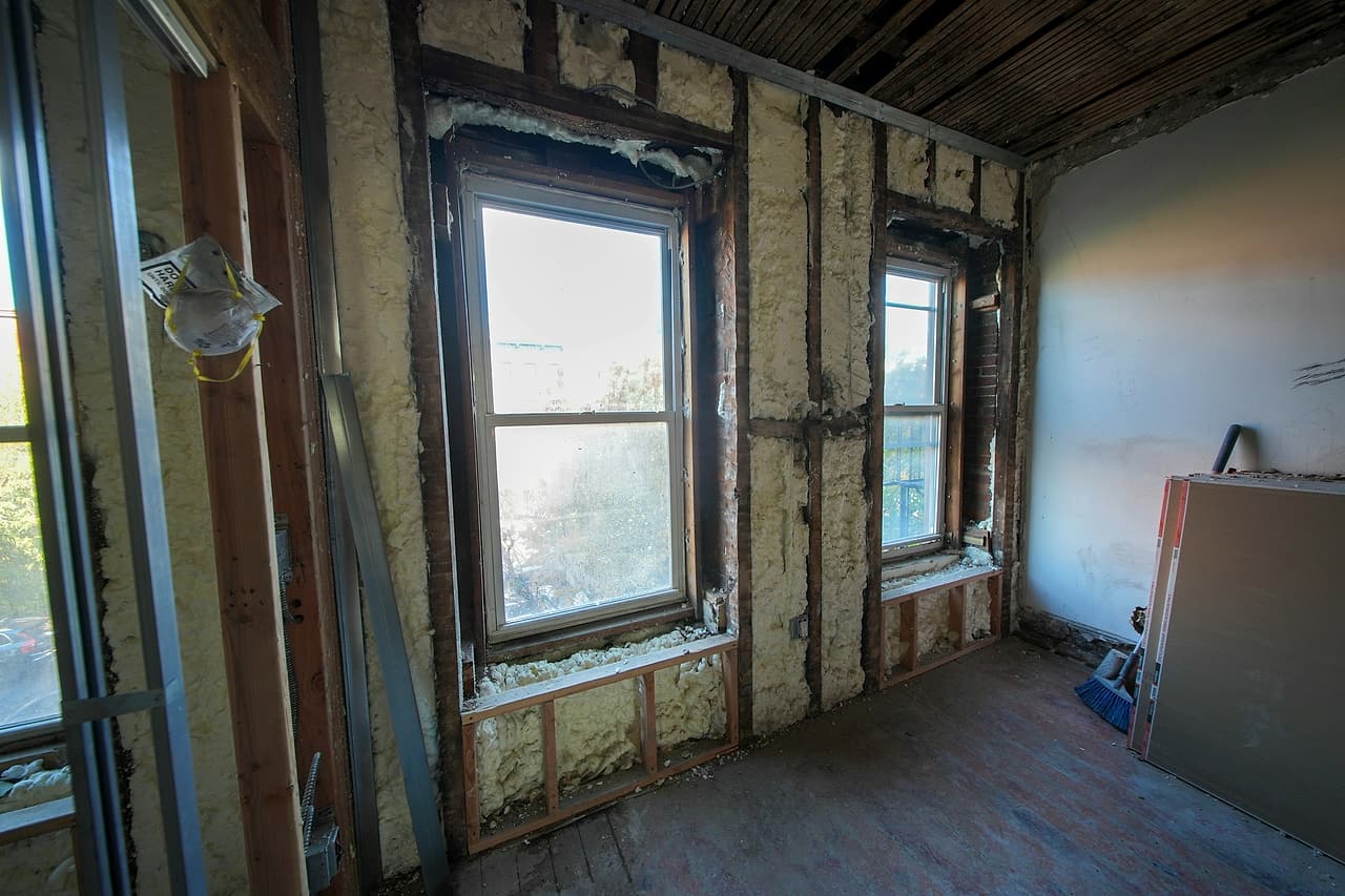 Exterior walls with spray foam between the studs around two windows, and drywall sheets leaning ready to close them in.