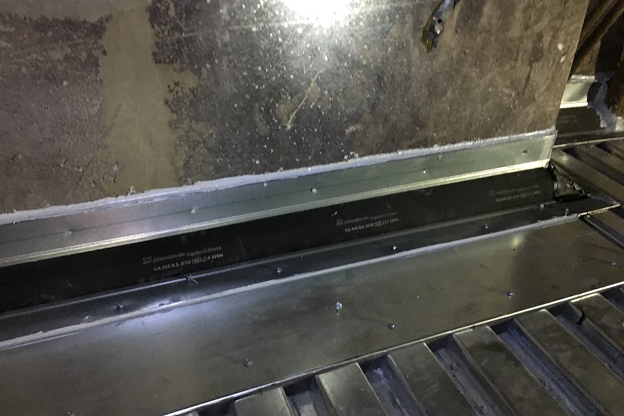 A metal flashing detail joins a concrete wall to corrugated decking, with a dark strip between metal termination pieces.