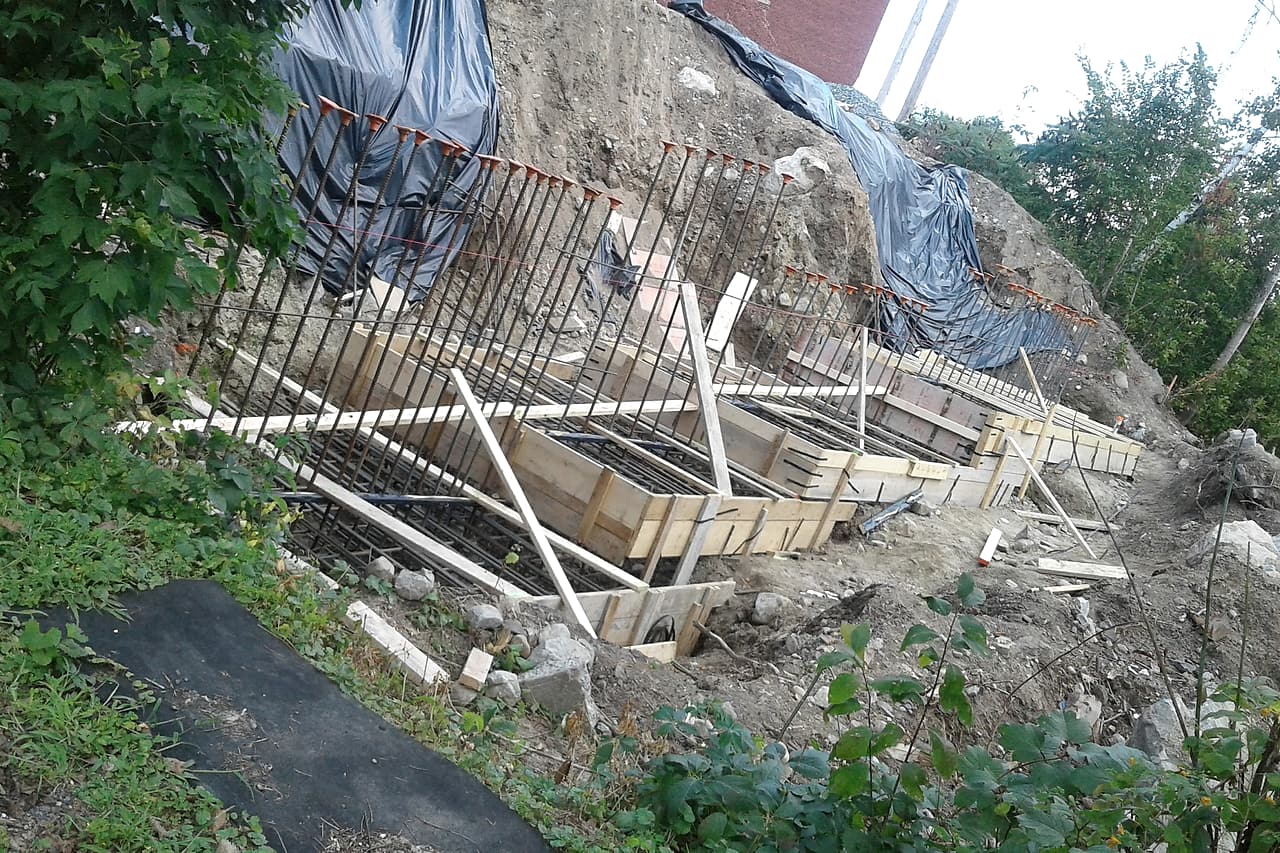 Timber footing forms and capped reinforcing bars follow an excavated bank, with black sheeting on the soil behind them.