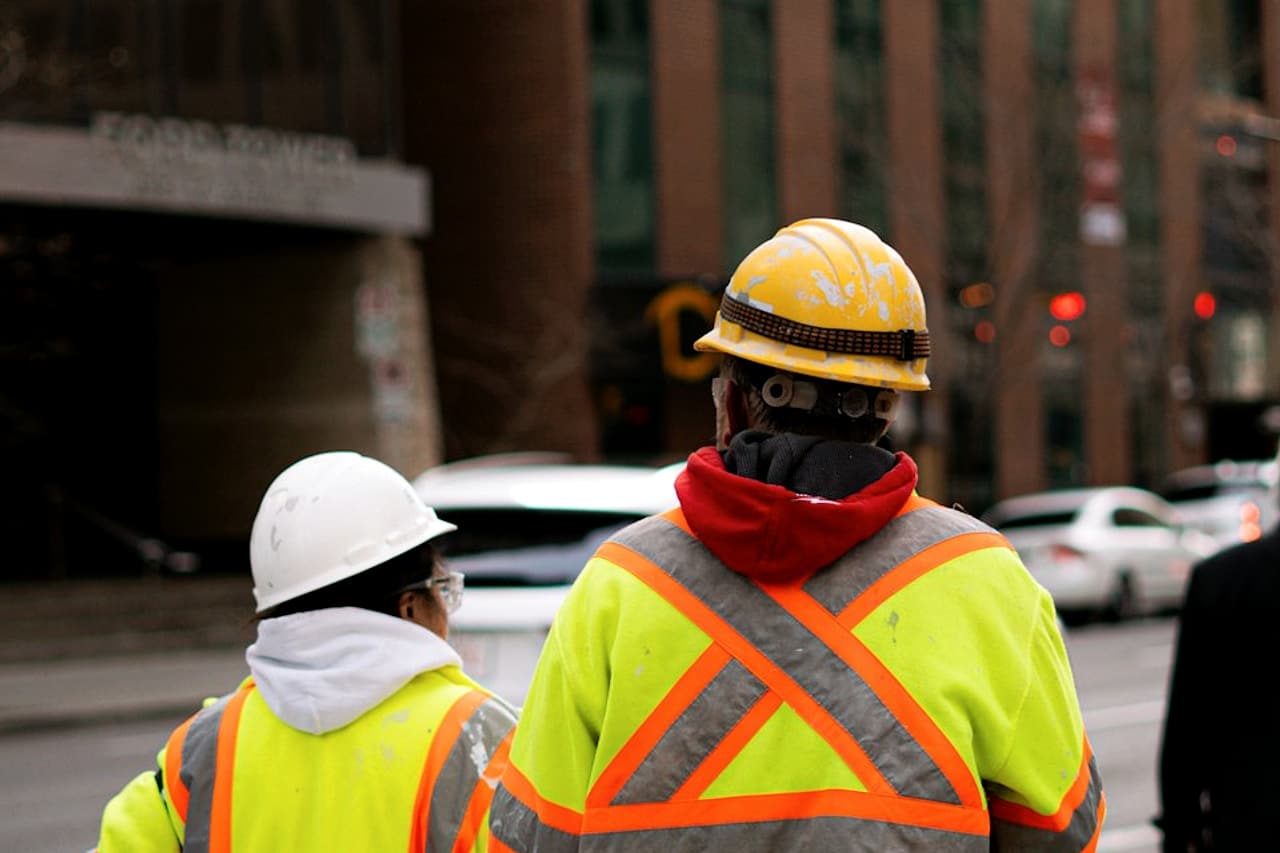 Two workers in hard hats and high-visibility clothing are seen from behind on a city sidewalk.