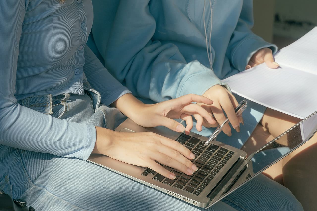 Two people use a laptop beside a blank lined notebook.