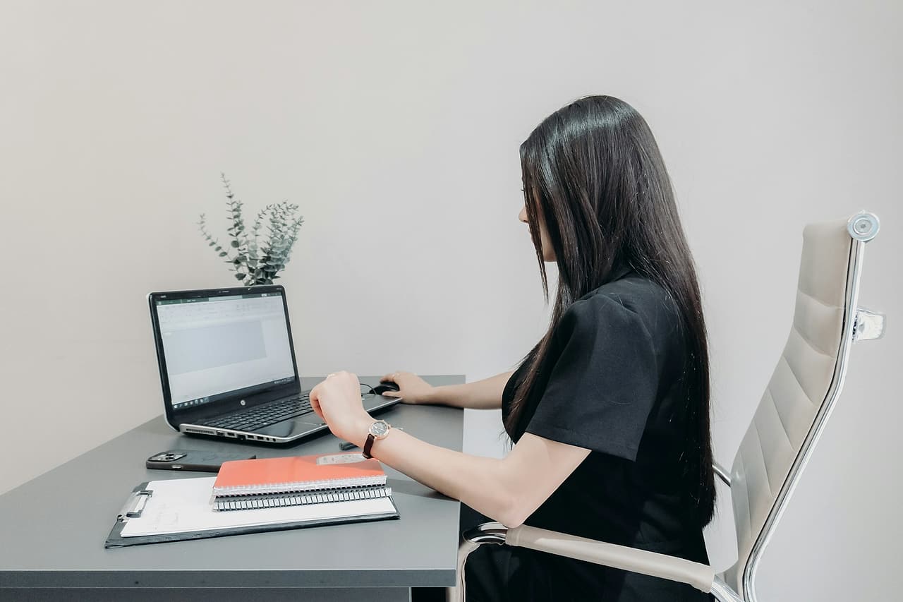 A person works at a laptop showing a spreadsheet-style document, with a clipboard and notebooks on a grey desk in a bright office.