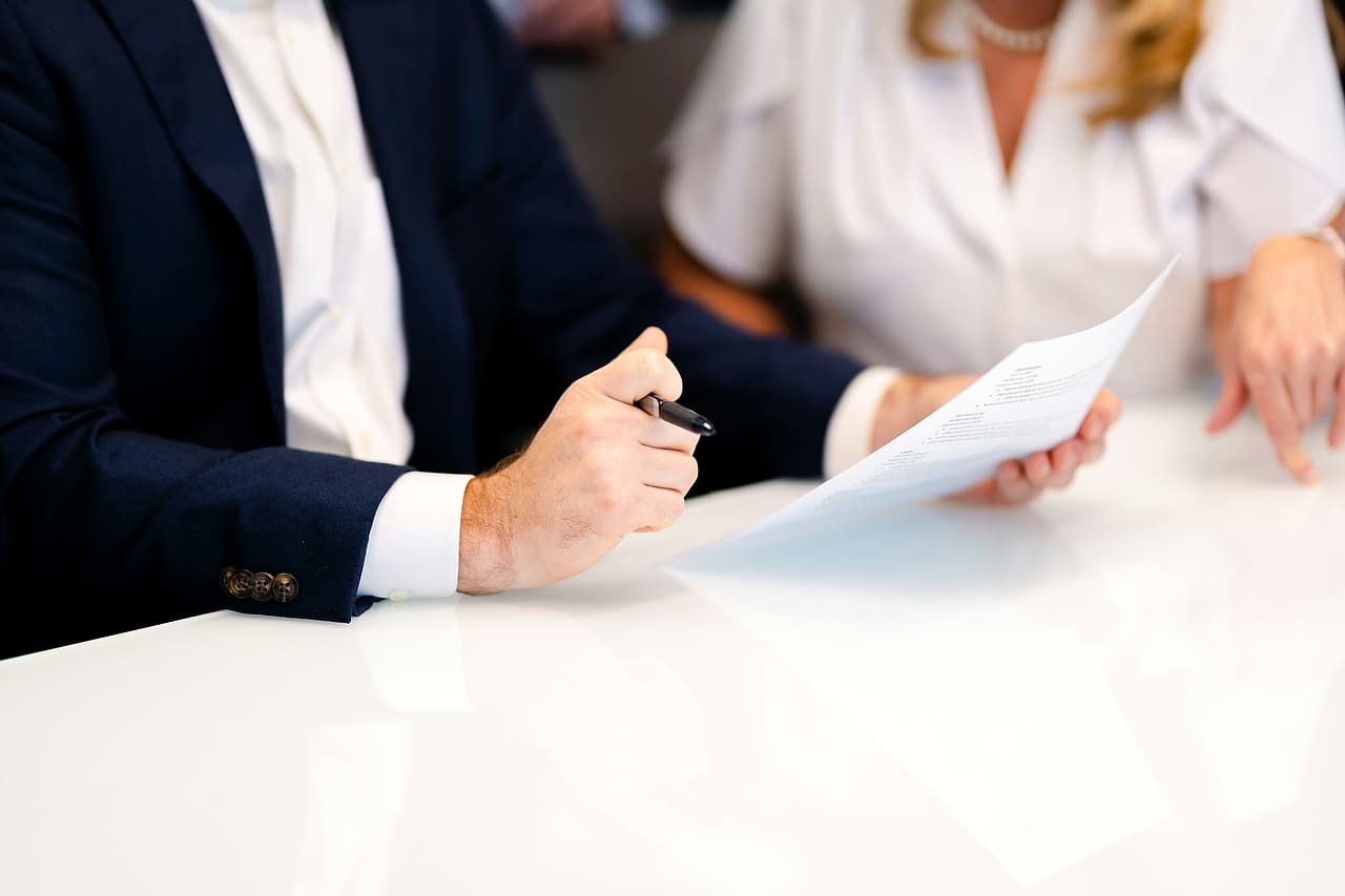 A man in a navy jacket holding a pen and a woman holding a document sit side by side at a white table.