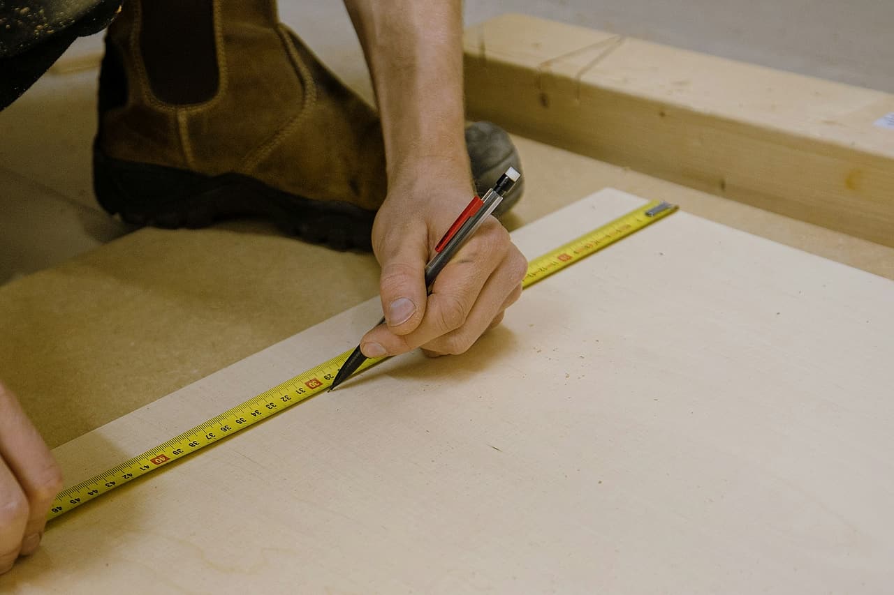 Hands measuring and marking a pale sheet of wood with a tape and pencil.