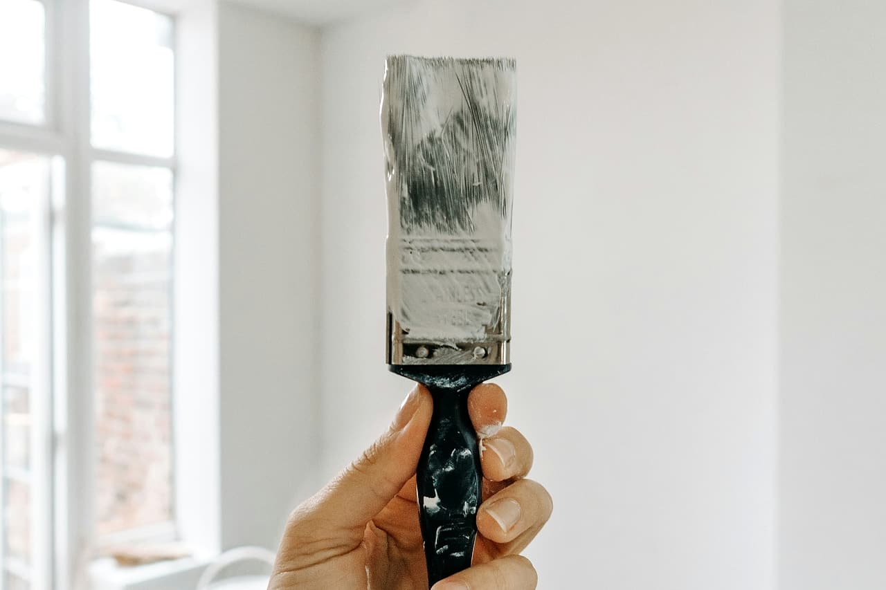 A hand holds a paint-covered brush in an unfinished room.