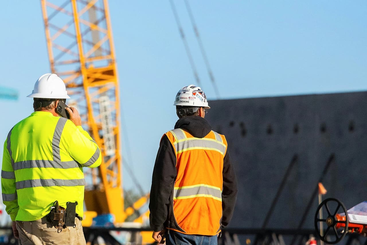 A worker in a hard hat and high-visibility shirt takes a phone call while a colleague watches a crane lift on site.