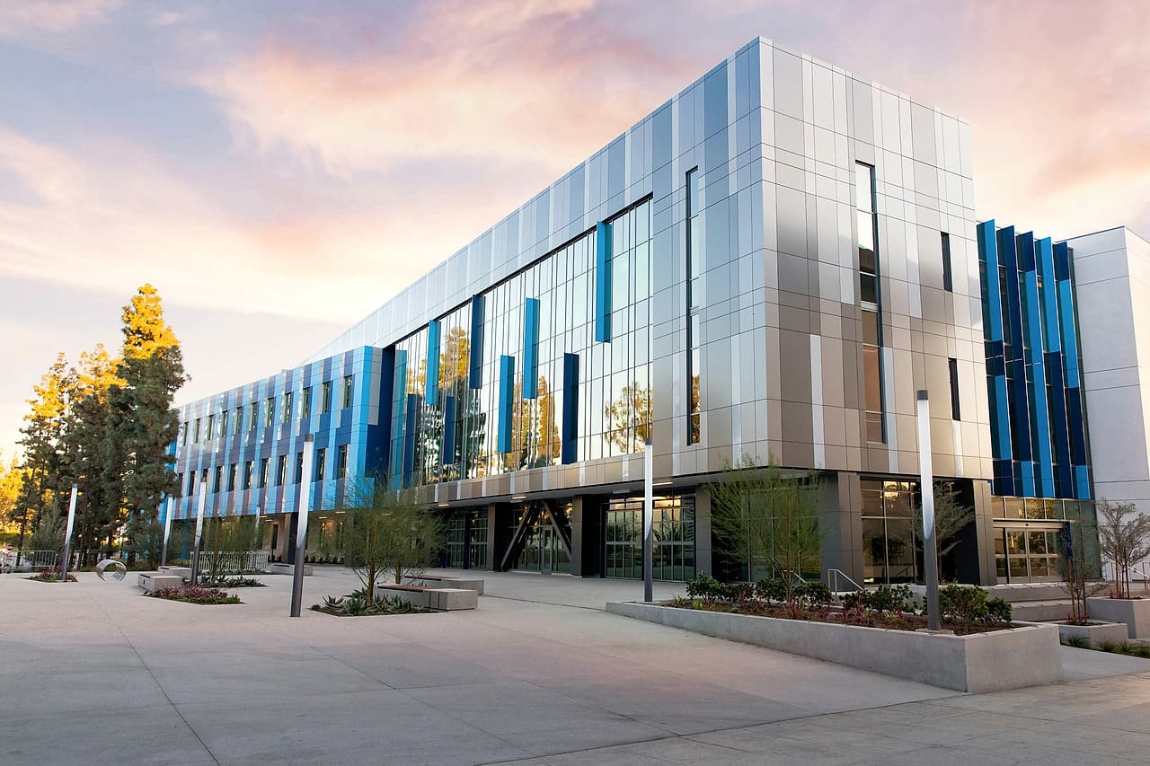 A newly completed three-storey building with blue and silver metal panels and a glass entrance, seen across its plaza at dusk.