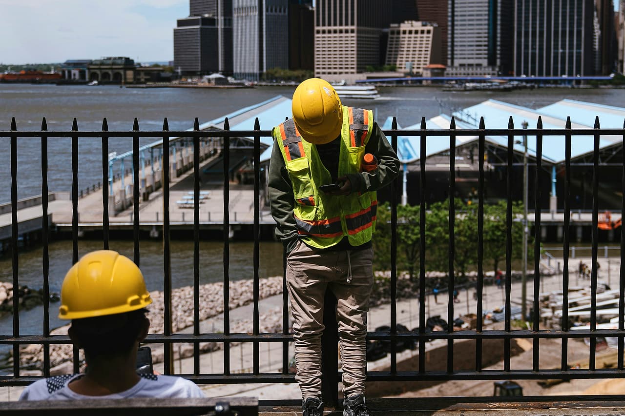 A worker in a hard hat and high-visibility vest reads his phone at a railing above a waterfront work site, with a city skyline across the river.