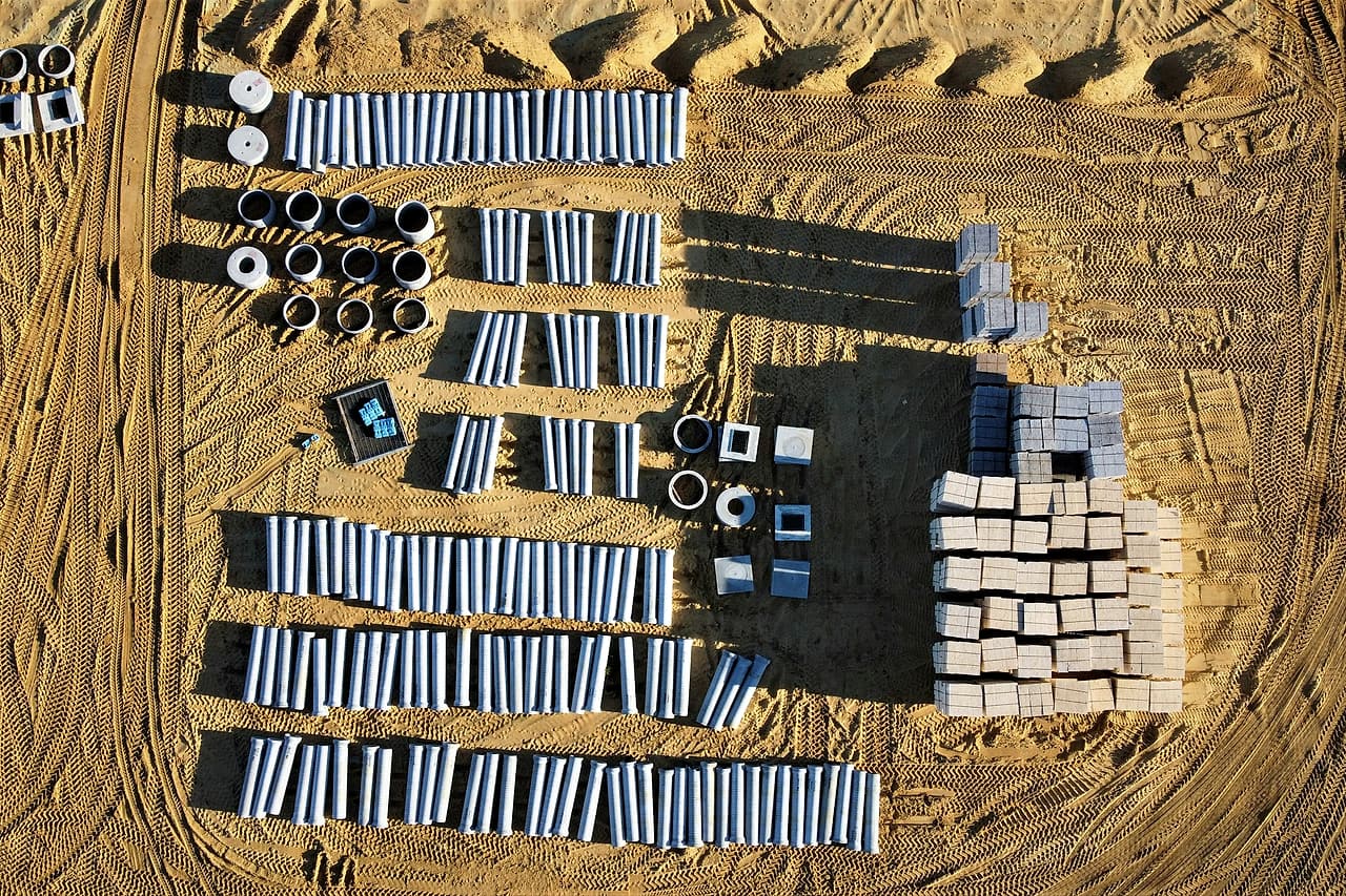 Seen from above, rows of pipe sections, manhole rings and precast blocks laid out on a graded site, waiting to be installed.