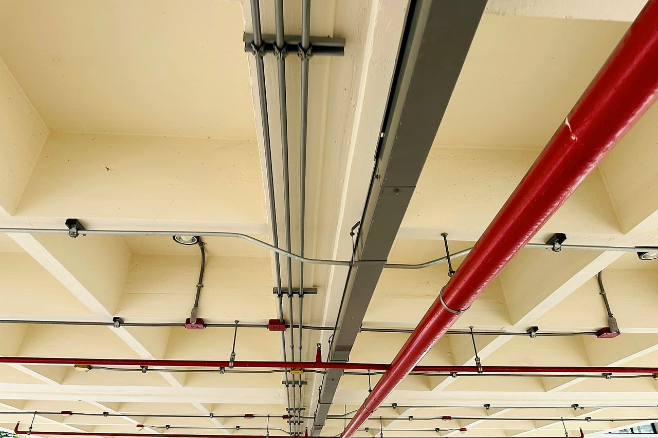 Steel conduits, a grey cable raceway and a red sprinkler pipe run side by side under a concrete waffle slab.