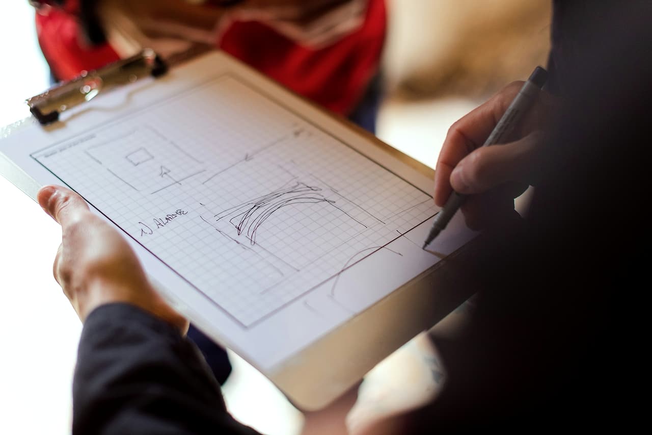 Hands hold a clipboard of grid paper while a pen marks up a sketched detail labelled with a numbered note.