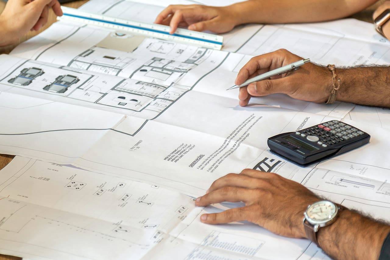 Three people's hands over printed house plans with a scale ruler, pencil and calculator on a shared table.