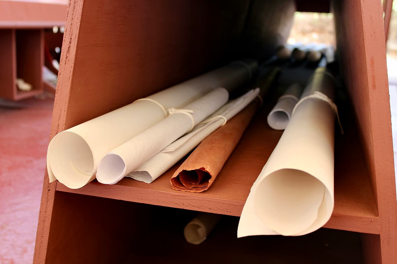 Several rolled drawing sets, some tied with string, lying together on the shelf of a wooden plan rack.