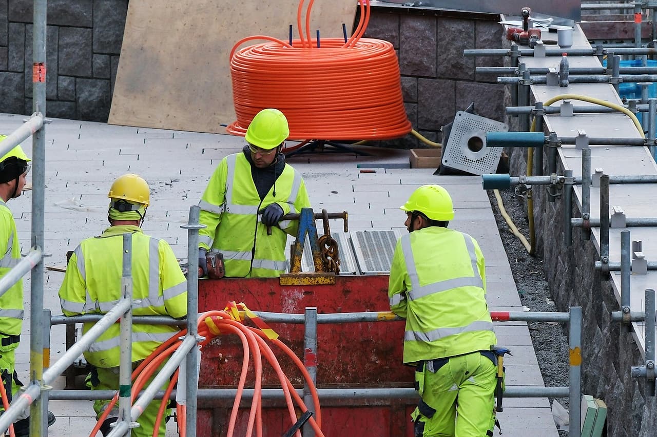 Four workers in hard hats and reflective jackets gather beside a work area surrounded by scaffold.