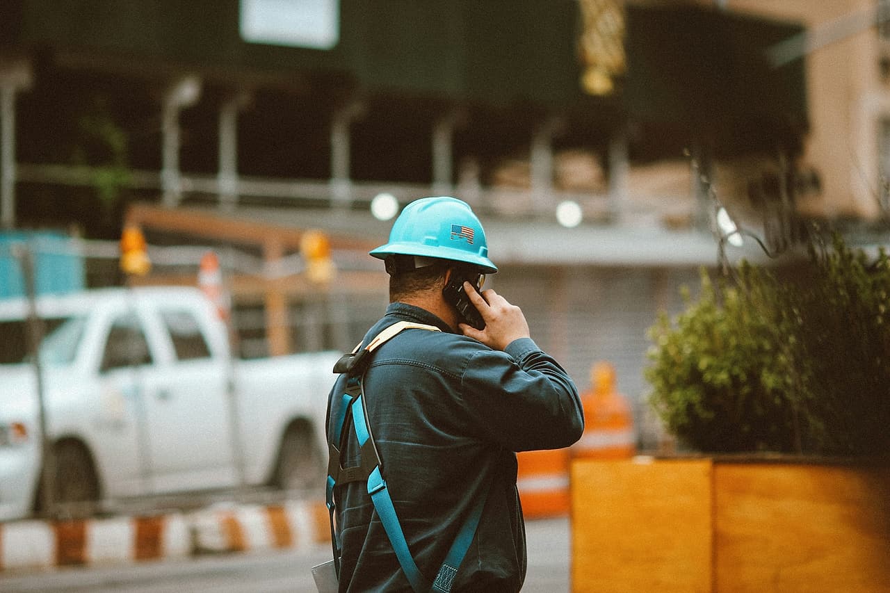 A worker in a hard hat makes a phone call beside a construction site.