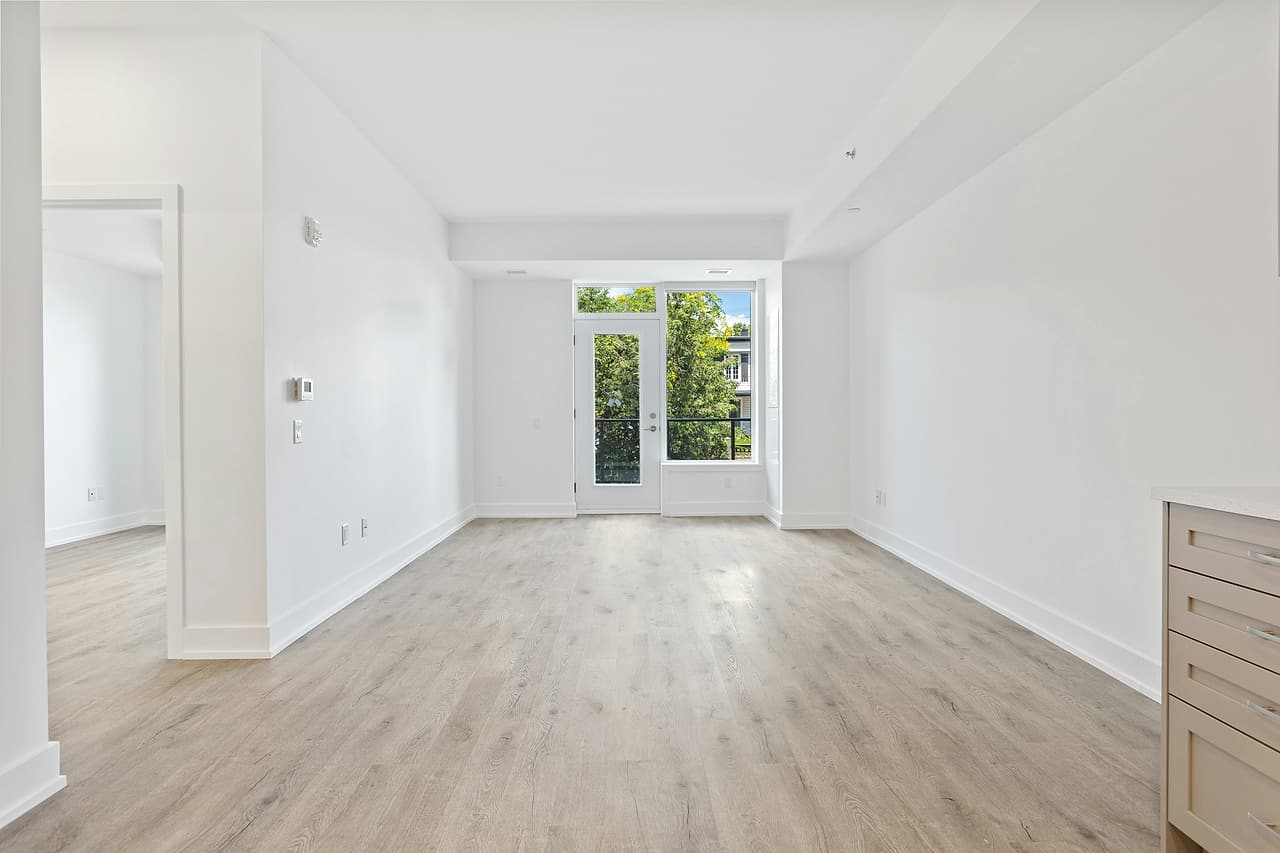 An empty, newly finished apartment with white walls, light plank flooring, kitchen cabinets and a glazed balcony door.