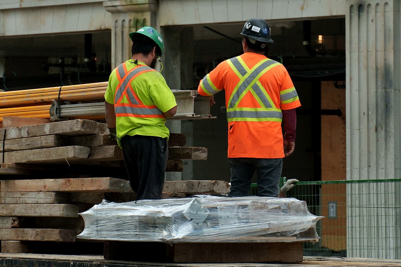 Two workers in hard hats and high-visibility shirts sort lumber on a flatbed truck parked beside a brick heritage building.