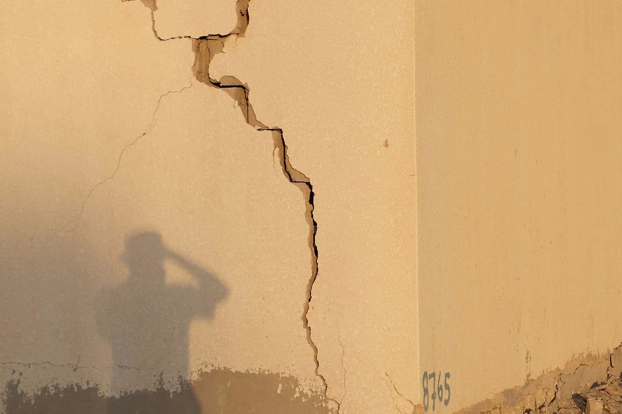 A deep crack runs down the corner of a stucco wall, with the shadow of a person photographing it.