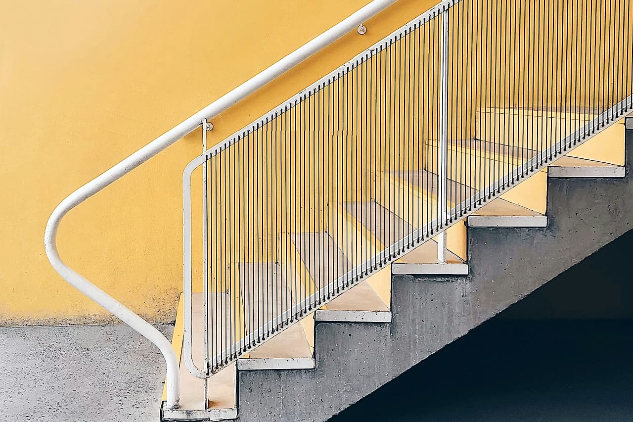 A white handrail follows an empty staircase beside a yellow wall.