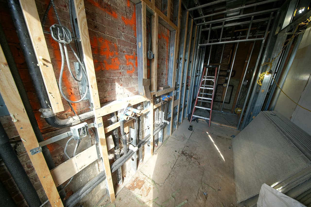 Open stud walls in a gutted brick building with capped plumbing stubs, electrical boxes and armoured cable before drywall.