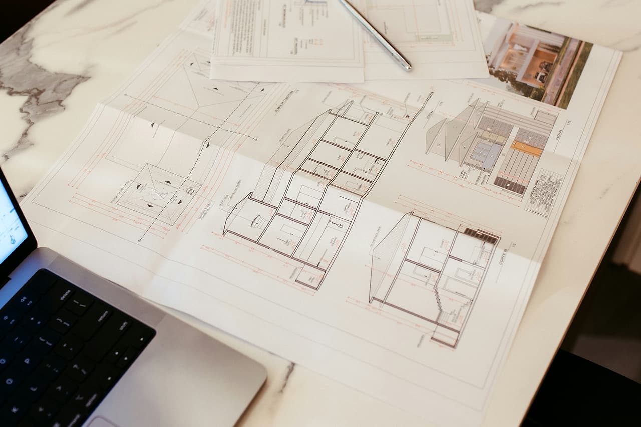 Architectural section drawings and a detail sheet spread on a desk beside an open laptop, a pen across them.