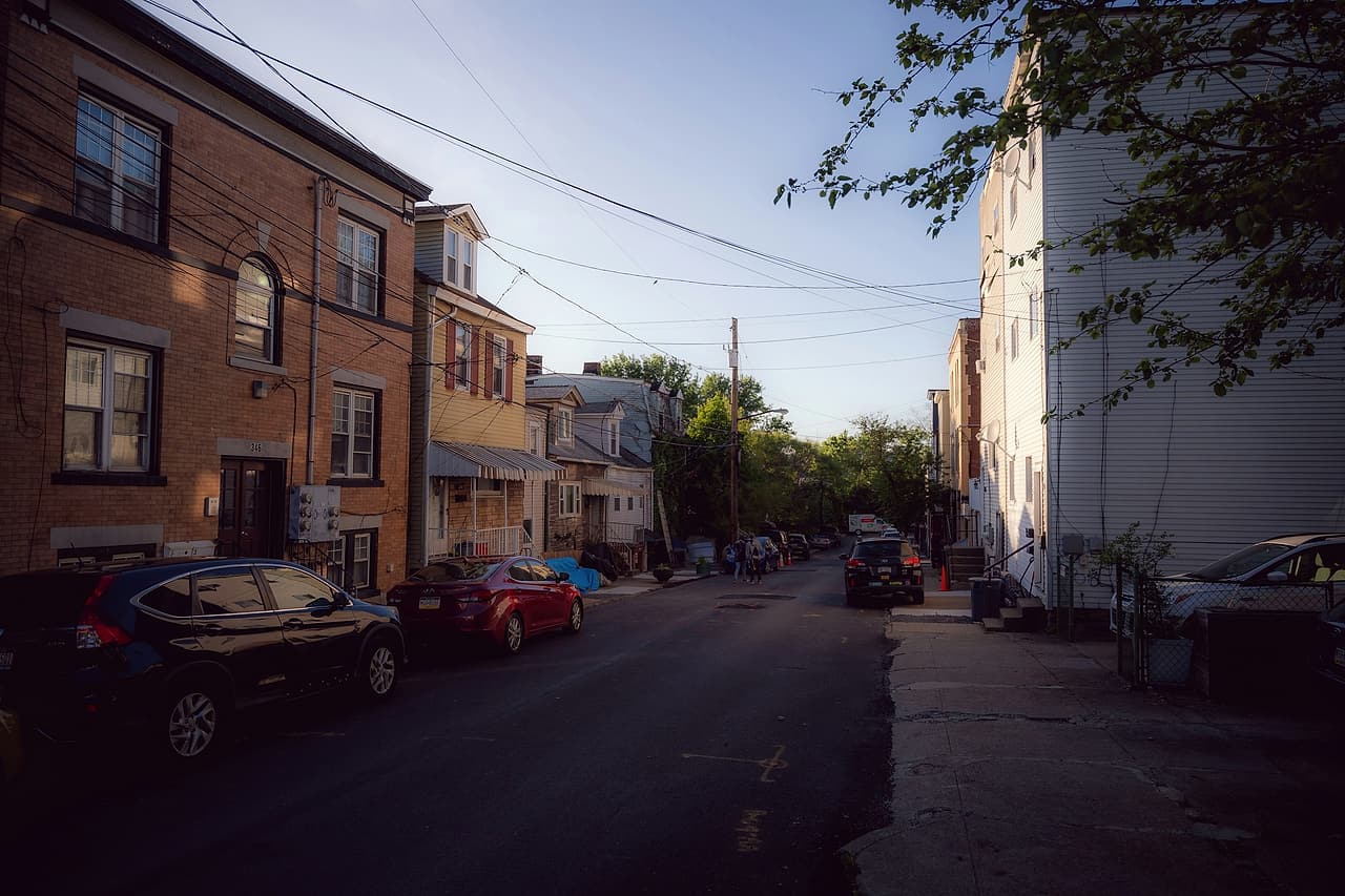 A narrow residential street of brick and sided row houses with parked cars, lit by low evening sun.