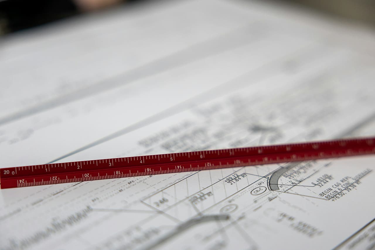 A red triangular engineer's scale lies across a civil drawing, its curb and grade notes in focus, the sheets behind soft.