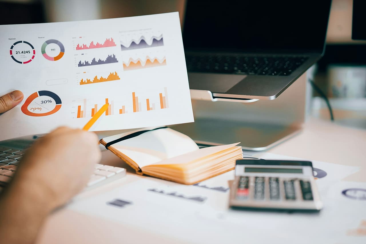 A hand points a pencil at a printed page of charts beside a laptop, an open notebook and a calculator.