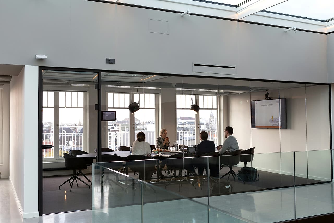 Four people meet around a table in a glass-walled meeting room, seen from the landing outside.