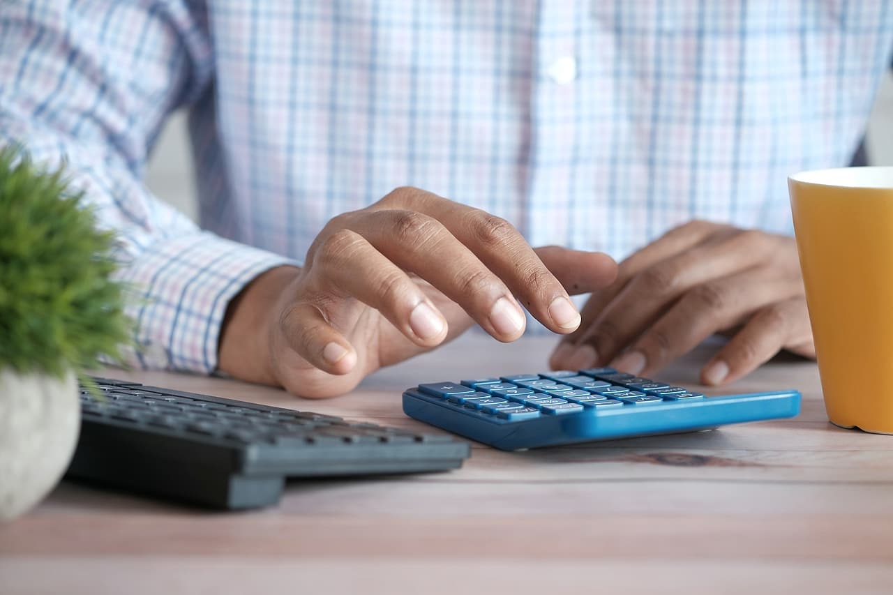 Hands use a blue calculator at a desk beside a keyboard and coffee mug.