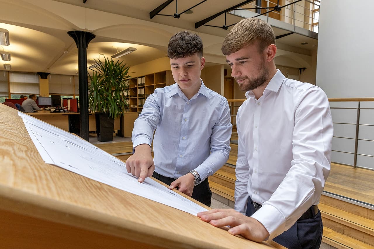Two people in an office review a large drawing on a sloped table, one pointing to a detail.