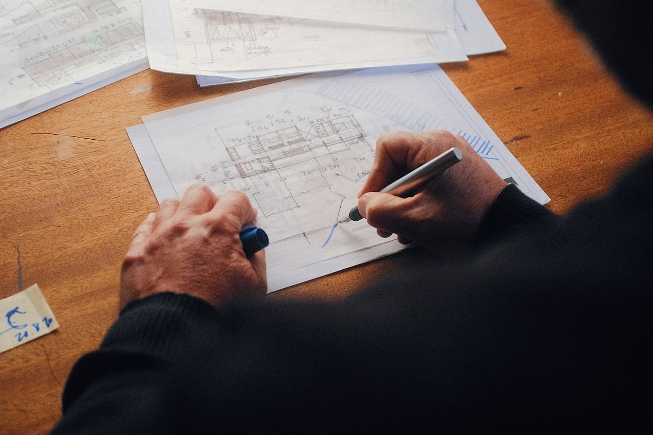 Seen over a shoulder, a person marks up floor plan sketches with a pen at a wooden table.