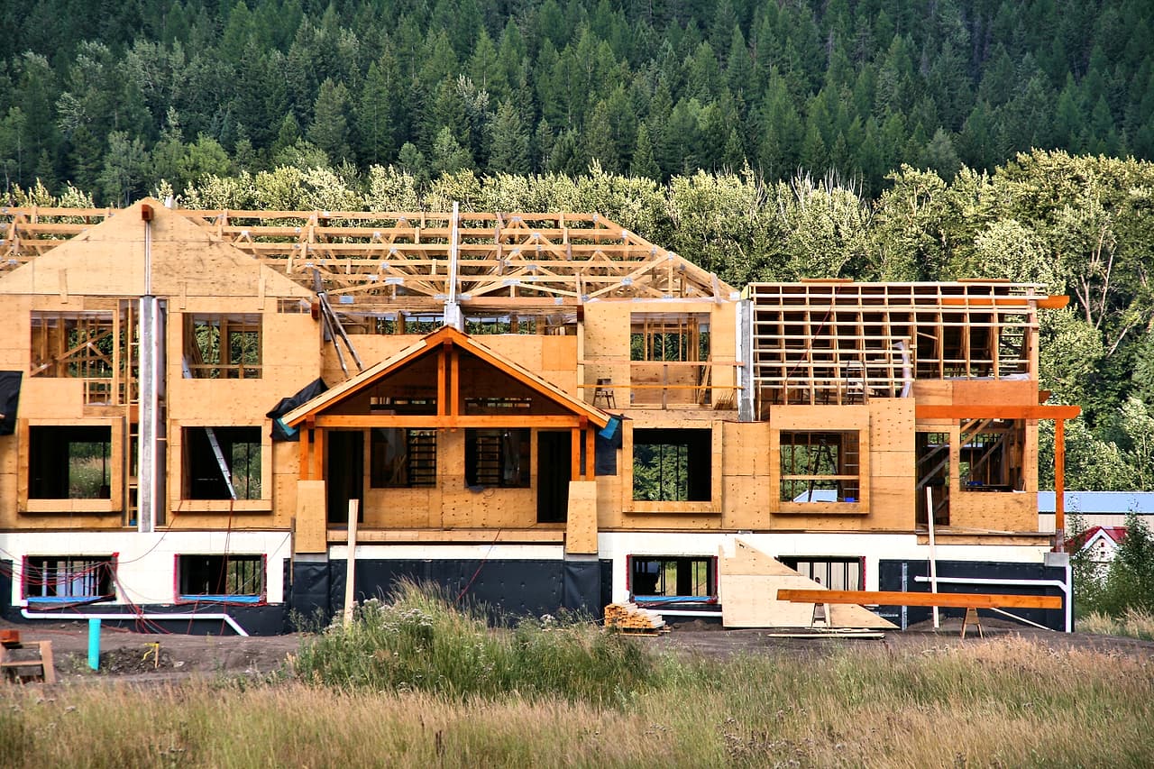 An unoccupied timber apartment building under construction shows repeated wall bays and roof framing against a forested hillside.