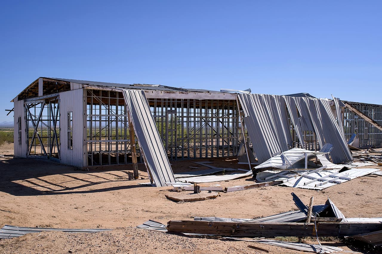 A steel-framed building with its metal cladding torn and bent, and sheet metal and lumber scattered on the ground around it.
