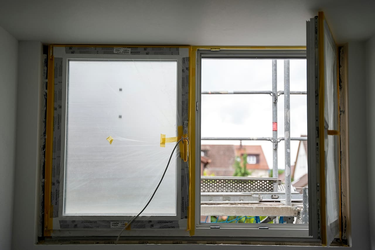 A newly installed window still in its protective film and tape, with a guarded scaffold visible through the open sash.