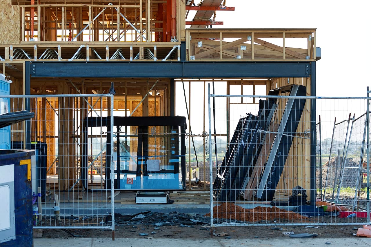 The timber frame of a building under construction, with a steel beam over its ground floor, behind temporary site fencing.