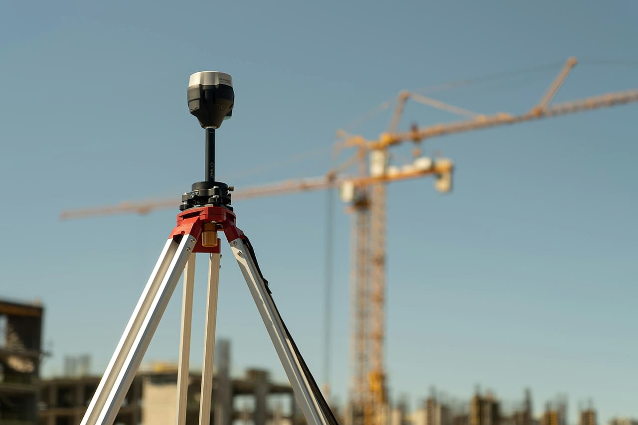 A GNSS receiver on a surveying tripod in front of a tower crane and concrete building frames.