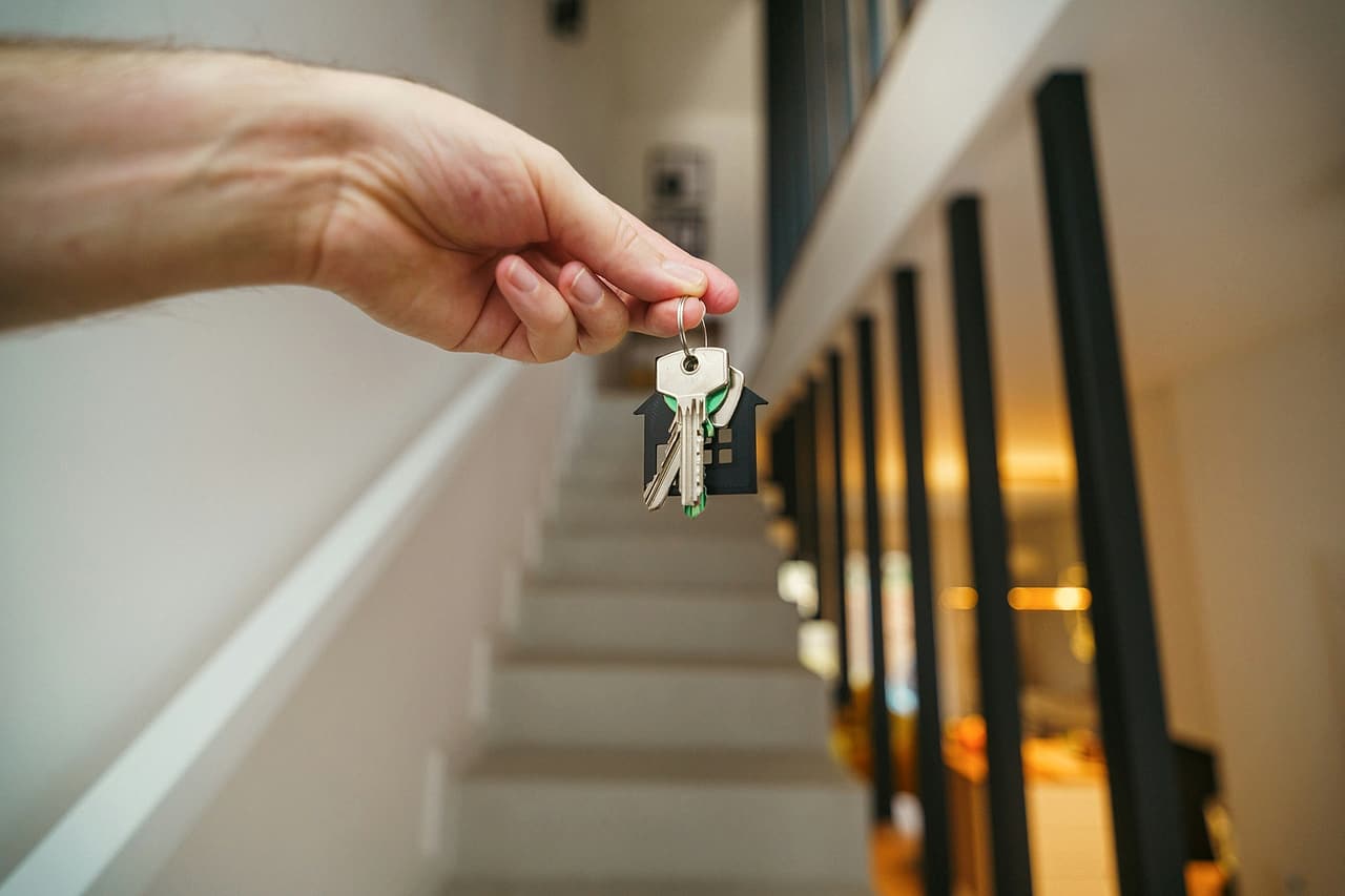 A hand holds out a set of keys on a house-shaped fob at the foot of a new staircase with black balusters.