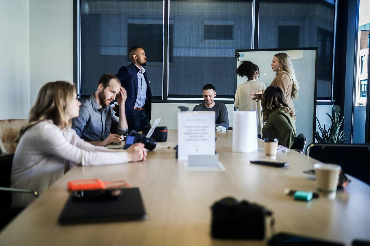 People around a conference table in a meeting, one man standing and another resting his head on his hand.