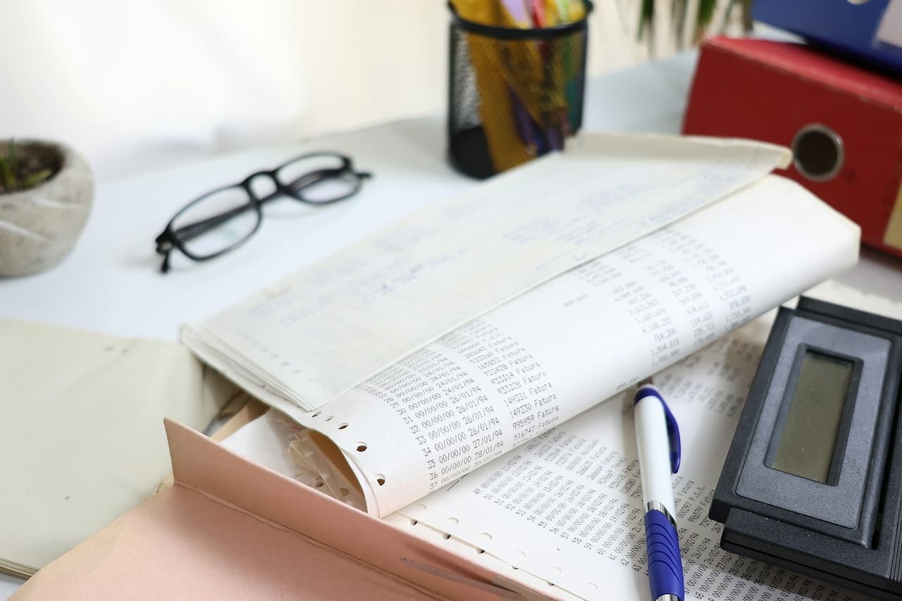 Printed invoice listings, a pen, a calculator, reading glasses and a red binder on an office desk.