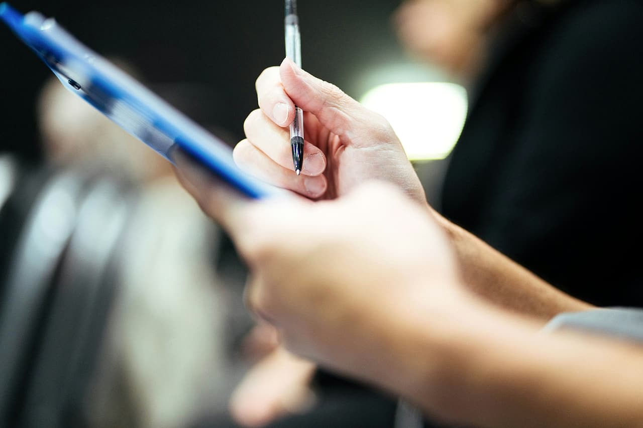A hand holding a pen over a blue clipboard, ready to fill in a checklist.
