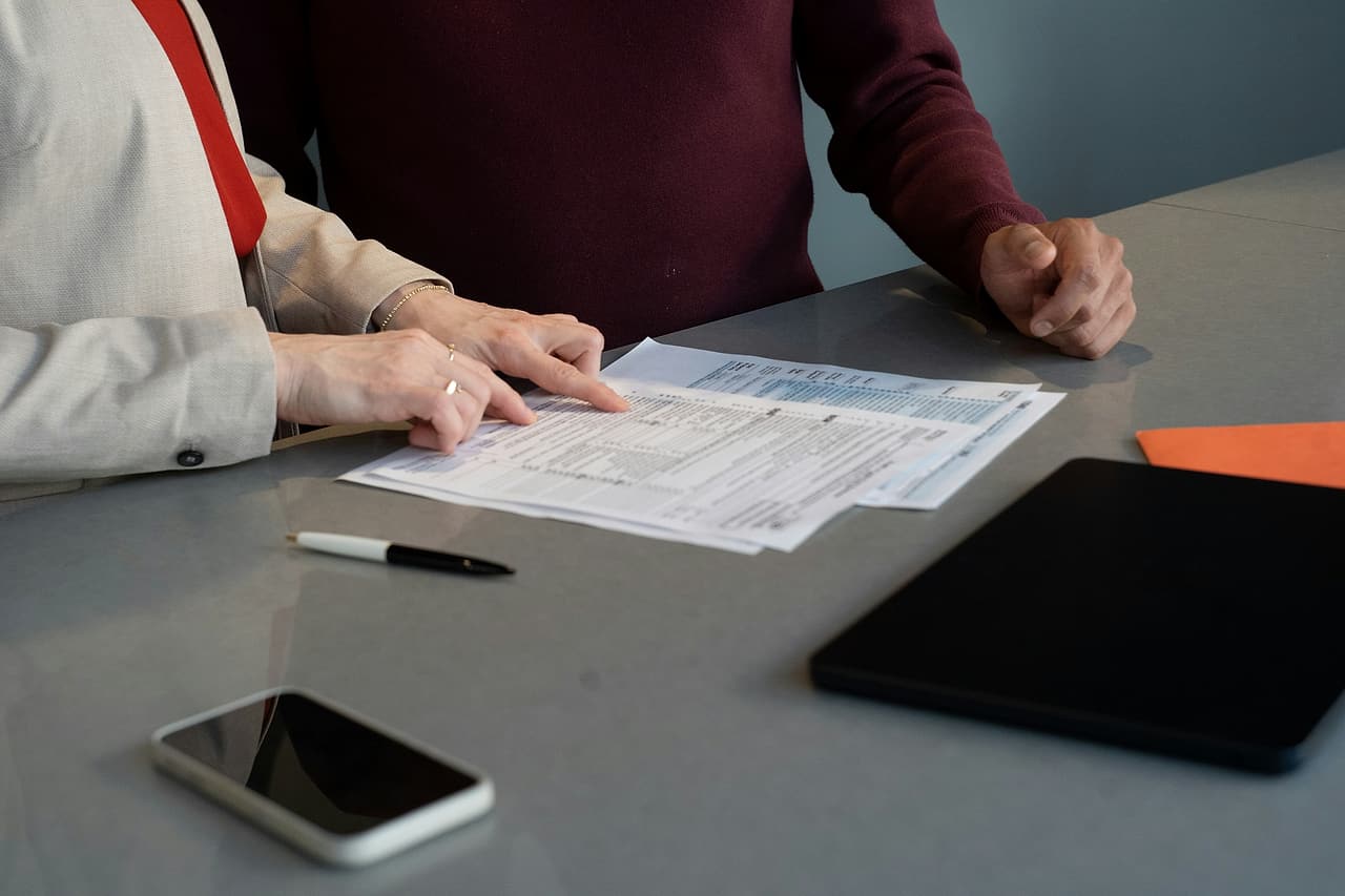 Two people at a grey table, one pointing at a line of a printed document, with a pen, a phone and a closed tablet beside it.