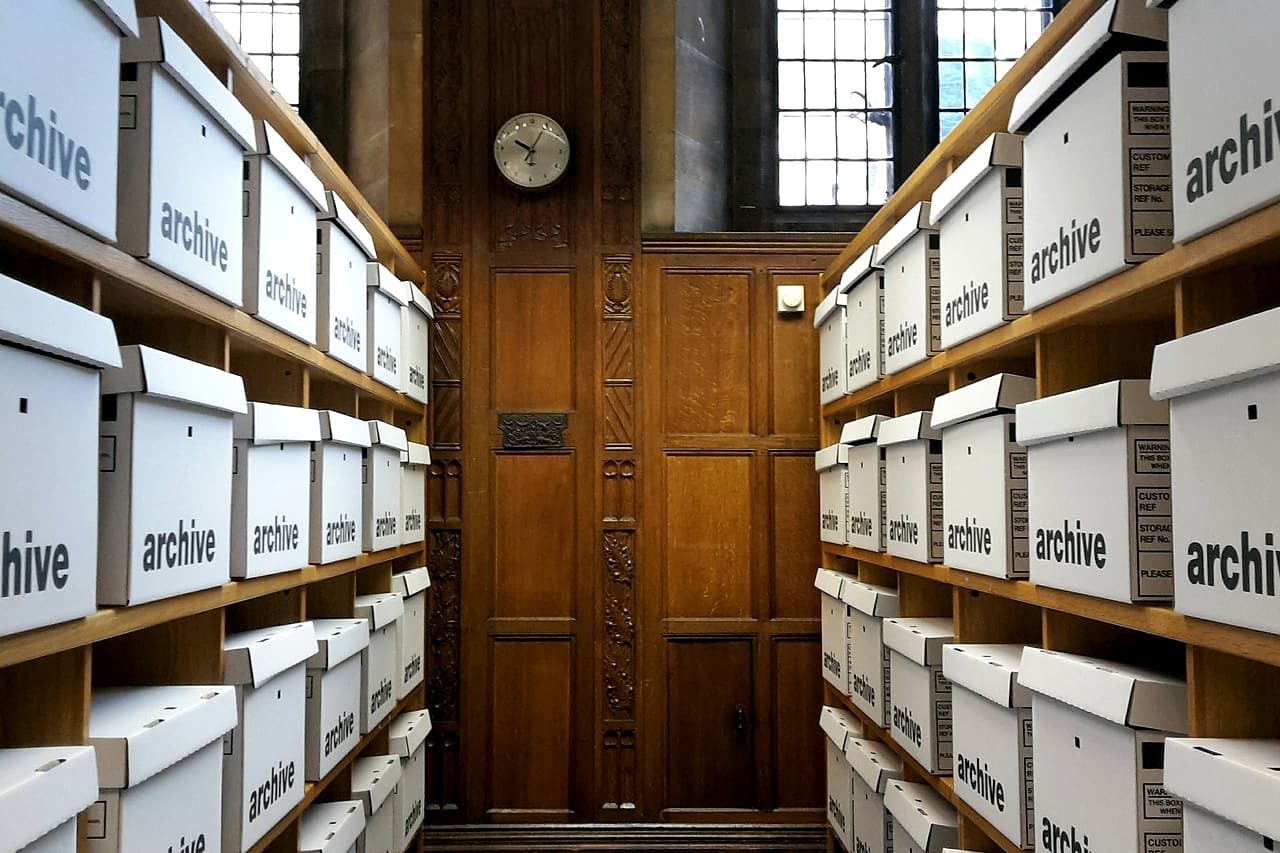 Rows of white archive boxes on wooden shelves line both sides of a panelled records room.