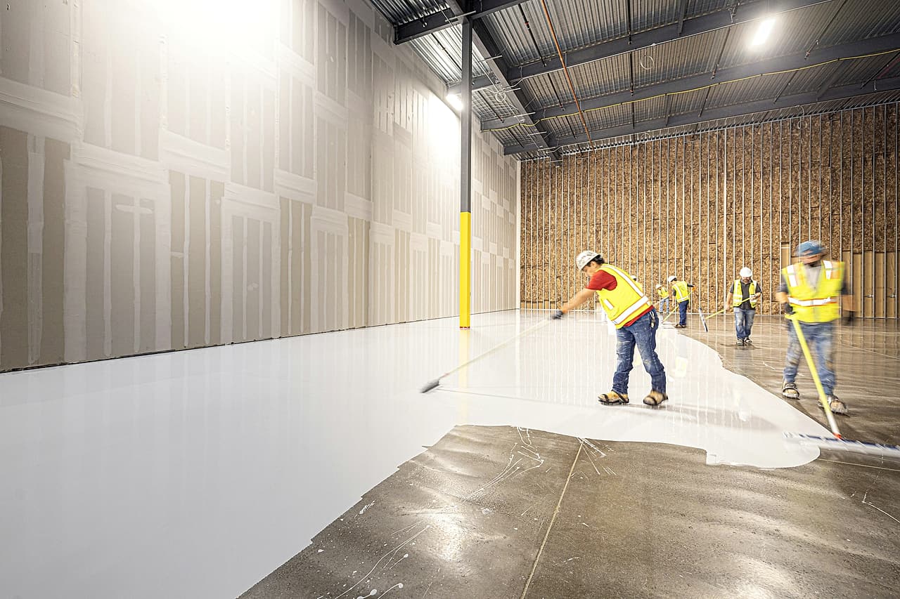 A crew in hard hats and high-visibility vests rolls a white coating across a warehouse floor, working in a line.