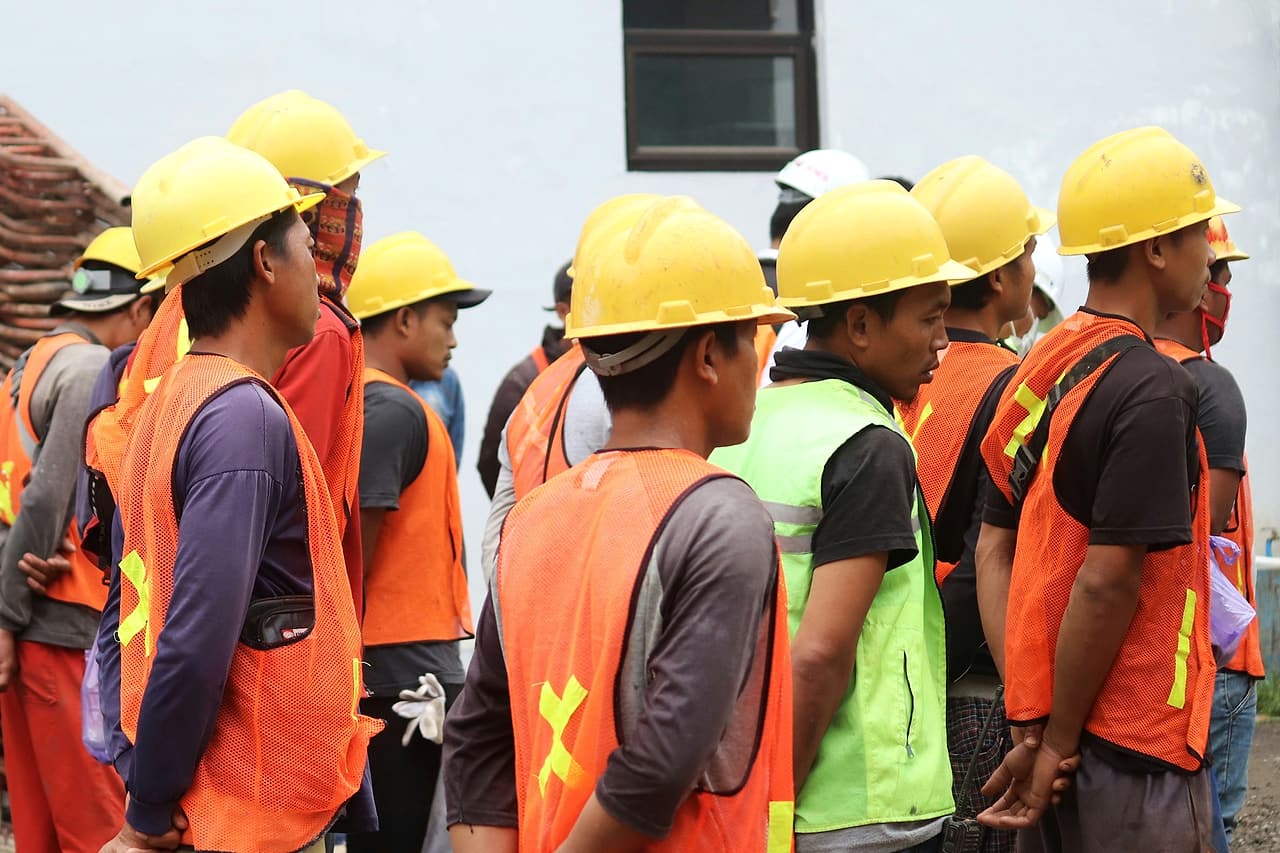 A crew in yellow hard hats and orange and green mesh vests stands listening during a site briefing.