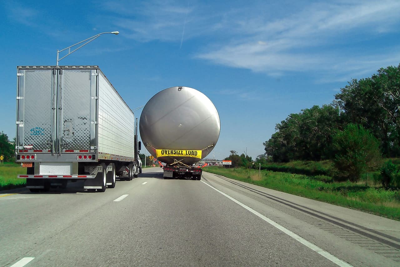 A large steel tank travels down a highway on a lowboy trailer under an Oversize Load banner, next to a transport truck.