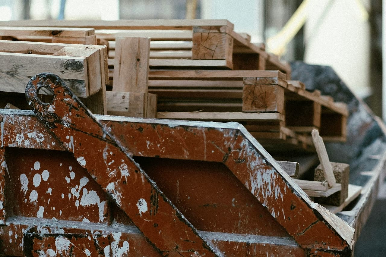Timber pieces sit in a worn metal hopper beside an unfinished building.
